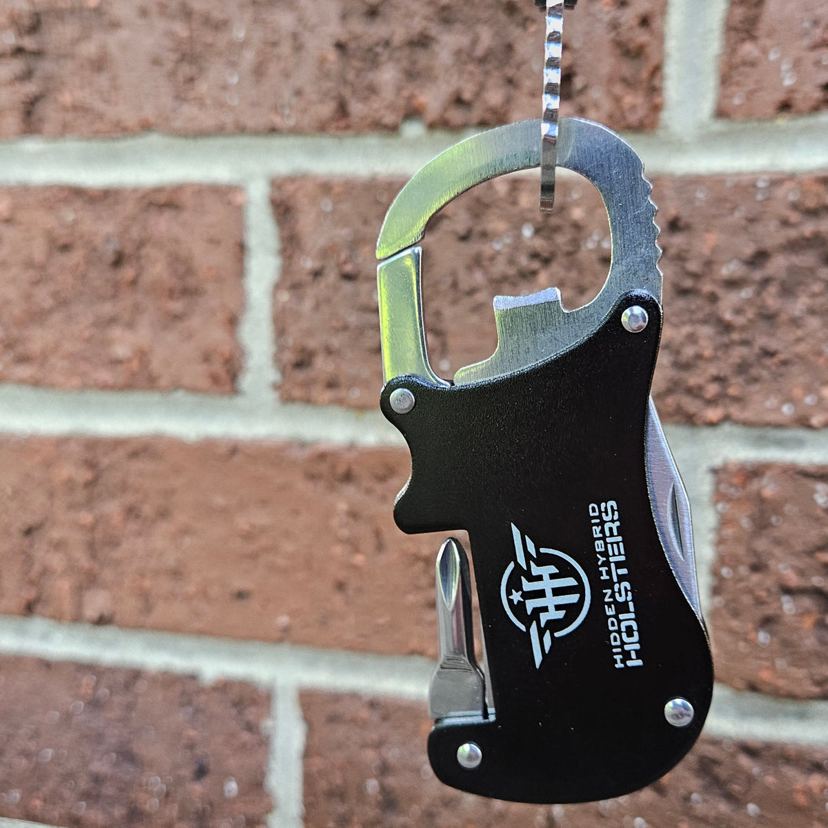 Metallic carabiner with bottle opener, philips screwdriver and letter opener on a wooden surface and brick wall background